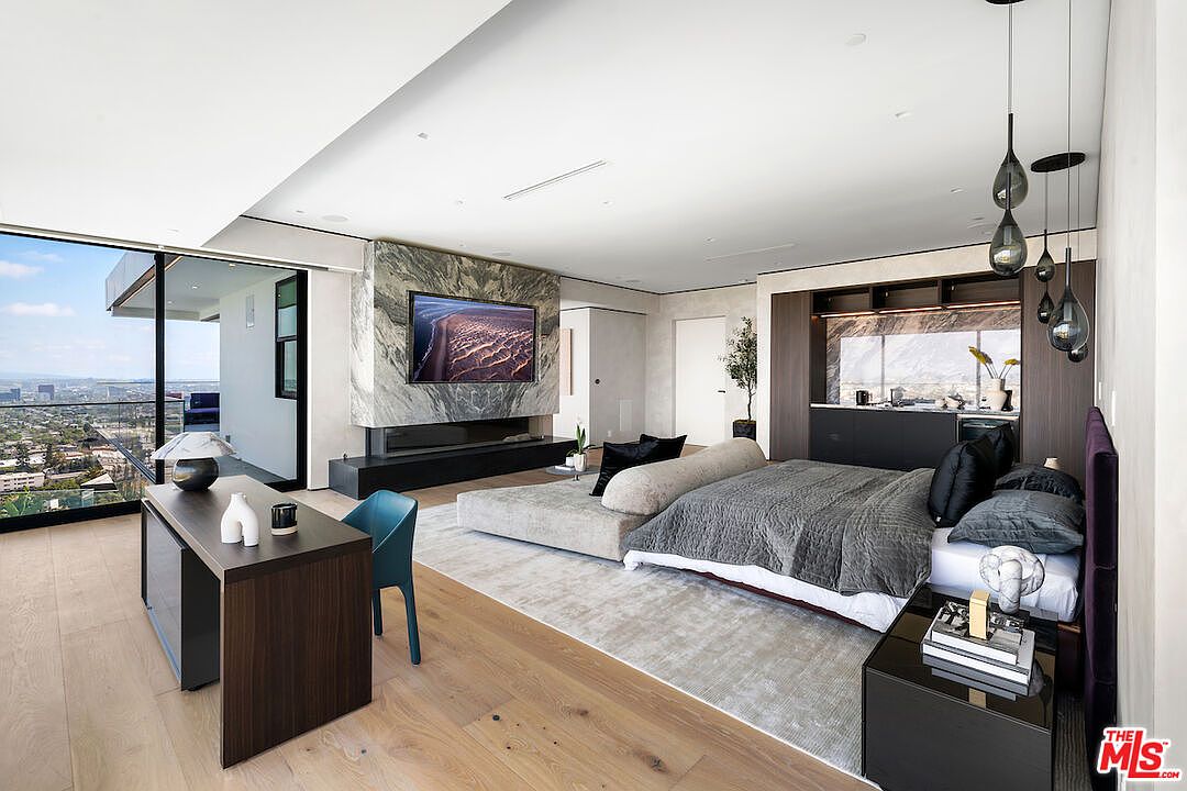 This expansive primary bedroom features a contemporary design with light wood flooring and a large area rug anchoring the sleeping space. A prominent stone-clad chimney breast serves as a focal point, housing a television and a linear fireplace, while a wall of glass provides panoramic city views. The room includes a dedicated workspace with a desk and a built-in wet bar area, all illuminated by modern pendant lighting.