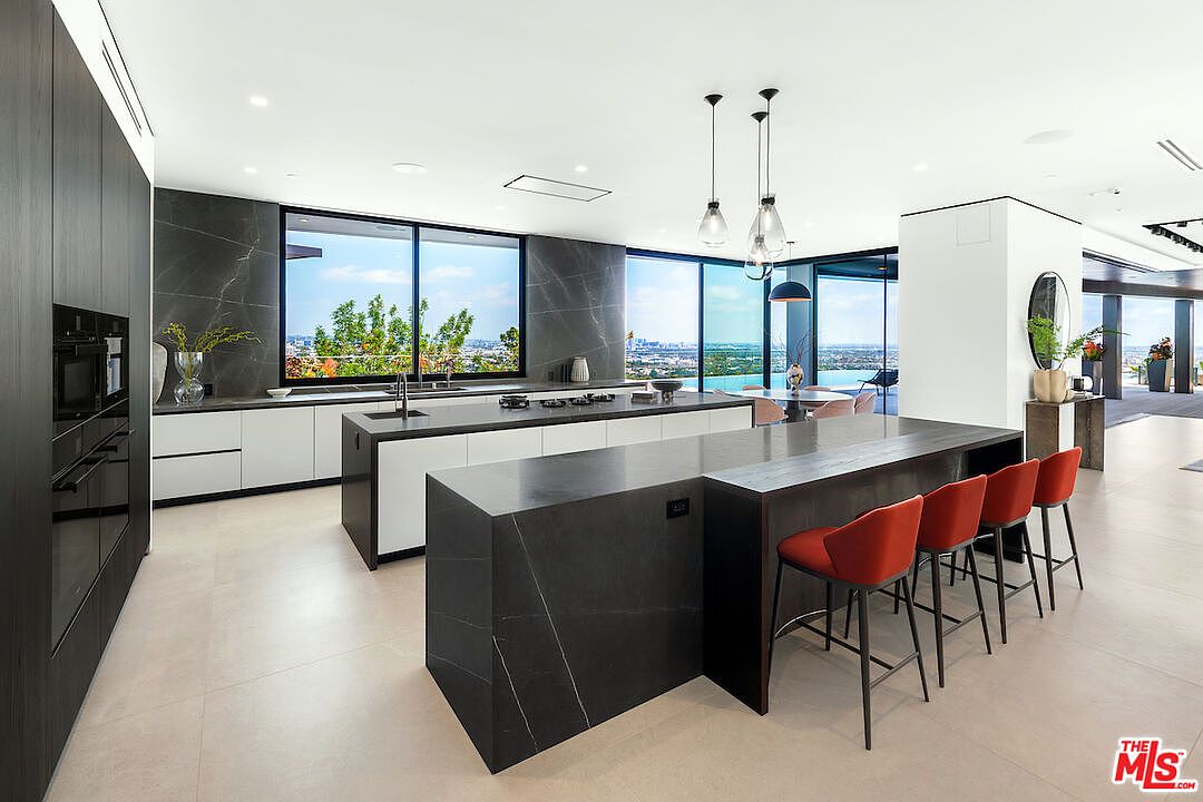 This ultra-modern, open-concept kitchen features a dual-island layout with dark stone surfaces and crisp white cabinetry. The space is defined by its expansive glass walls that provide panoramic views, complemented by minimalist pendant lighting and vibrant red bar stools. The sleek, high-contrast design creates a sophisticated and airy atmosphere perfect for luxury entertaining.