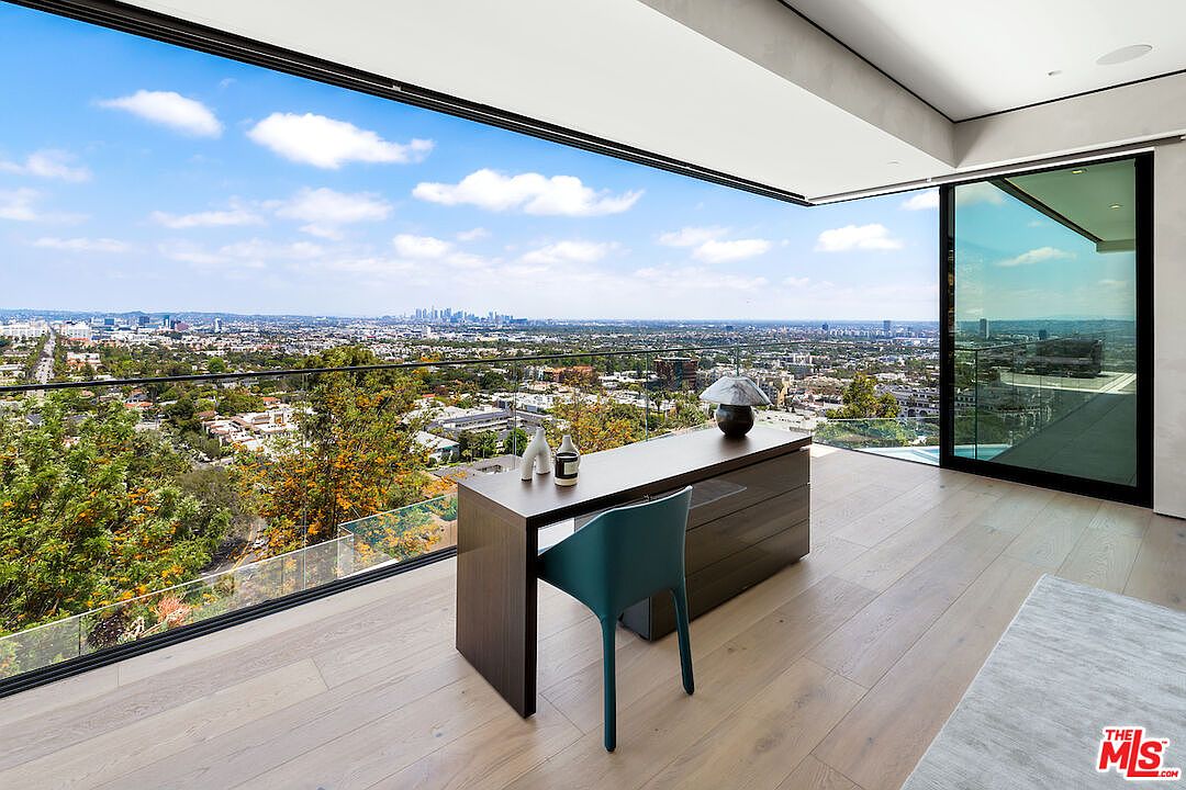 This modern office space features a minimalist dark wood desk positioned to face a panoramic view of the city skyline. The room is enclosed by expansive floor-to-ceiling glass walls that provide an unobstructed perspective of the urban landscape and surrounding greenery. The light wood flooring and sleek teal chair create a sophisticated, airy atmosphere perfect for a high-end home workspace.