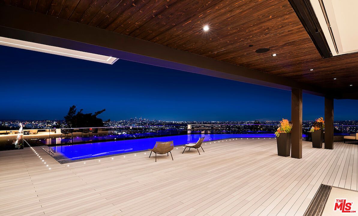 This expansive, covered outdoor deck features light-toned decking and a sleek, modern infinity-edge pool that overlooks a sprawling city skyline at night. The space is sheltered by a dark wood-paneled ceiling with recessed lighting, creating a sophisticated and luxurious atmosphere perfect for evening entertaining. Two lounge chairs are positioned poolside, emphasizing the seamless transition between the indoor-outdoor living space and the panoramic urban views.