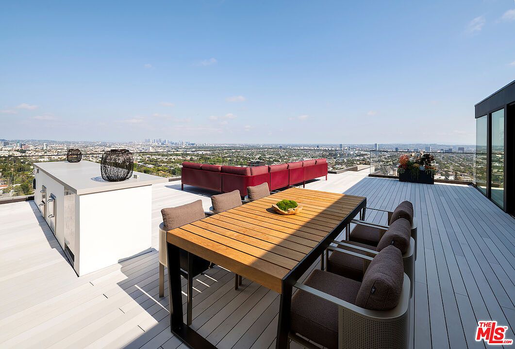 This expansive rooftop deck offers a sophisticated outdoor living space with a panoramic view of the city skyline. The area is furnished with a long wooden dining table surrounded by cushioned chairs and a large, deep-red sectional sofa, complemented by a sleek outdoor kitchen island. The clean, modern aesthetic is enhanced by light-toned decking and a glass railing that ensures an unobstructed view of the urban landscape.