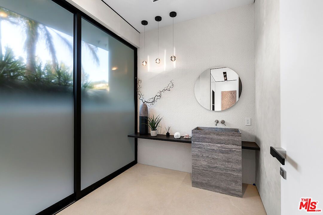 This modern powder room features a striking stone pedestal sink and a minimalist floating shelf against a textured white wall. A large circular mirror hangs above the sink, complemented by three sleek pendant lights, while a large frosted glass sliding door provides privacy and soft, diffused natural light. The space is designed with a clean, contemporary aesthetic that emphasizes geometric lines and high-end materials.