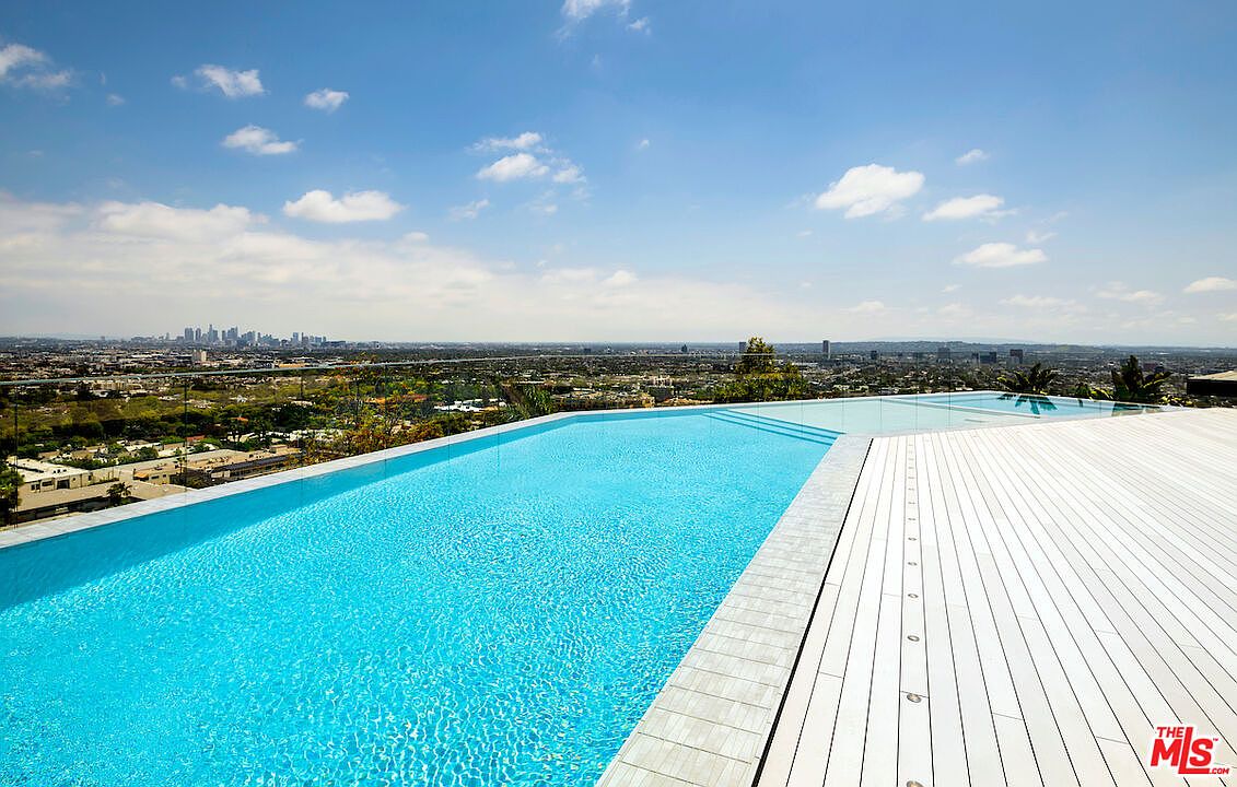 This image captures a stunning infinity-edge pool that seamlessly blends with the expansive city skyline in the distance. A wide, light-toned wooden deck borders the pool, providing a sleek space for lounging while overlooking the urban landscape. The perspective is elevated, emphasizing the dramatic drop-off of the pool edge and the panoramic view under a bright, partly cloudy sky.