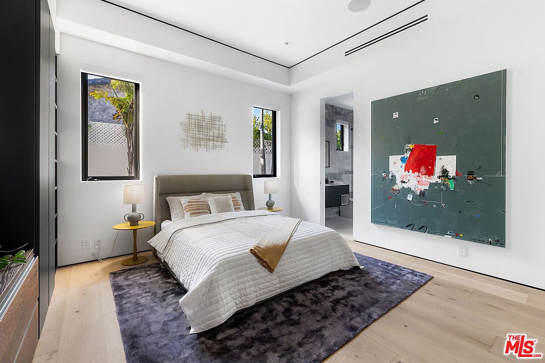 This contemporary bedroom features light wood flooring, a neutral-toned upholstered bed, and a large, abstract art piece mounted on the wall. Two tall windows flank the bed, providing natural light, while a dark, textured area rug adds depth to the minimalist space. The room includes a view into an adjacent bathroom, showcasing a clean, modern aesthetic.