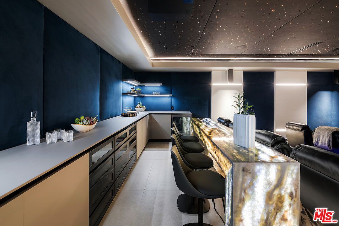 This sophisticated home bar and lounge area features a long, illuminated stone-topped bar with modern stools, set against a backdrop of deep blue velvet-textured walls. The ceiling is designed with a starry night effect, complemented by recessed lighting and sleek, minimalist cabinetry along the back wall. The space is arranged for entertainment, with comfortable theater-style seating visible in the background, creating an upscale, cinematic atmosphere.