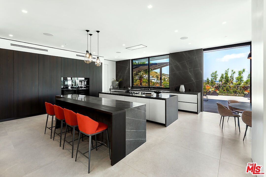 This sleek, modern kitchen features a large central island with a dark stone countertop and four vibrant orange bar stools. The space is defined by dark cabinetry, a matching dark stone backsplash, and a secondary island that houses the cooktop. Large glass doors open to an outdoor deck, creating a seamless transition between the indoor cooking area and the scenic exterior view.