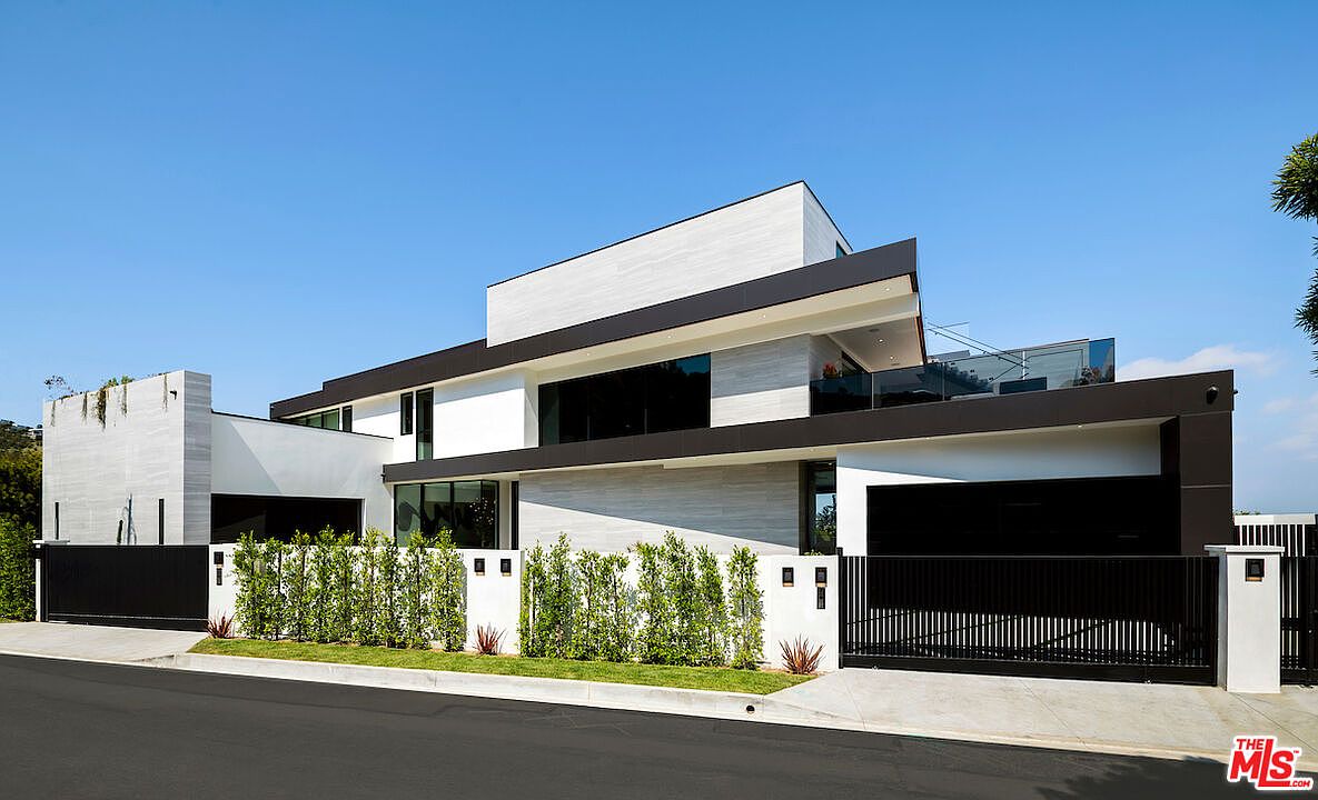 This modern, multi-level residence showcases a striking architectural design with clean lines, contrasting white and dark horizontal accents, and expansive glass surfaces. The property is secured by a sleek, dark metal gate and a white perimeter wall fronted by a manicured hedge, creating a private and sophisticated street presence. The perspective captures the home from the street level, highlighting its contemporary aesthetic and the integration of indoor-outdoor living spaces on the upper terrace.