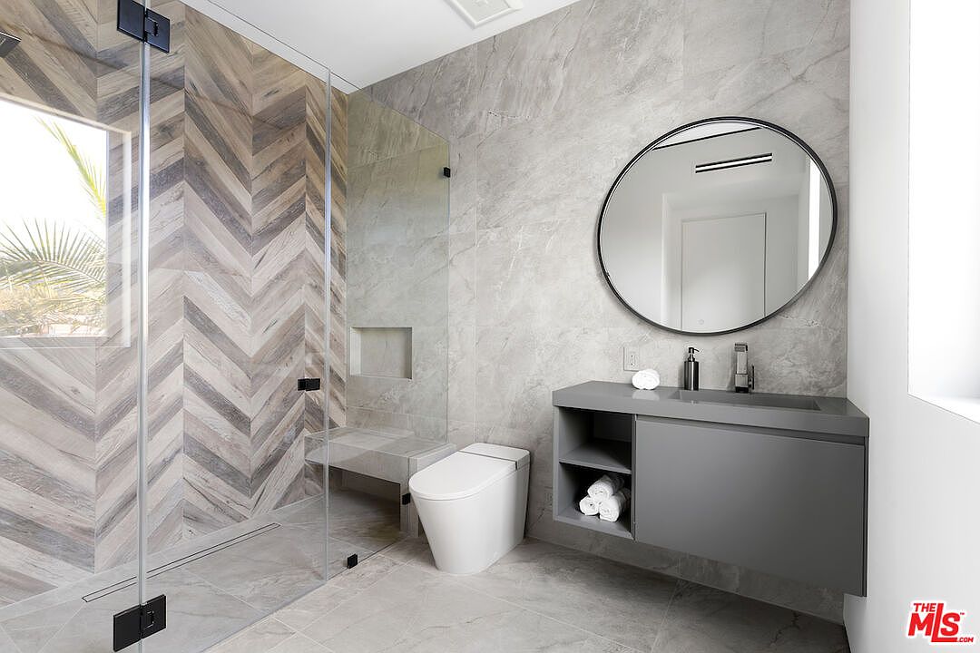 This modern bathroom features a walk-in shower with a striking chevron-patterned tile accent wall and a glass enclosure. A floating gray vanity with an integrated sink and a circular mirror sits against a large-format stone wall, complemented by a sleek white toilet. The space is bright and contemporary, utilizing a neutral color palette and clean lines to create a sophisticated, spa-like atmosphere.
