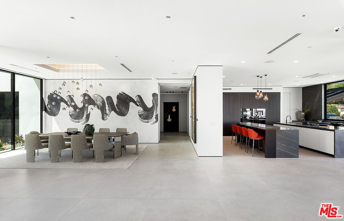 This expansive, open-concept living space features a dining area on the left with a large table and modern chairs set against a striking, abstract black-and-white mural. To the right, a sleek kitchen area includes a long island with red bar stools and dark cabinetry, creating a sophisticated and contemporary aesthetic. The room is illuminated by minimalist pendant lighting and recessed ceiling lights, with large glass panels providing a seamless connection to the outdoors.
