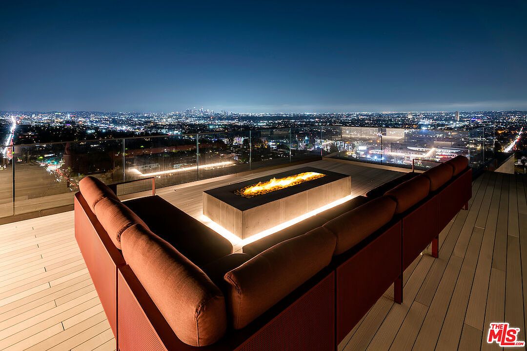 This elevated outdoor deck features a modern, rectangular fire pit as its centerpiece, surrounded by a plush, L-shaped sectional sofa with warm-toned cushions. The space is enclosed by a sleek glass railing that provides an unobstructed, panoramic view of the sprawling city lights at night. The perspective is captured from behind the seating area, emphasizing the luxurious, open-air atmosphere and the dramatic urban backdrop.