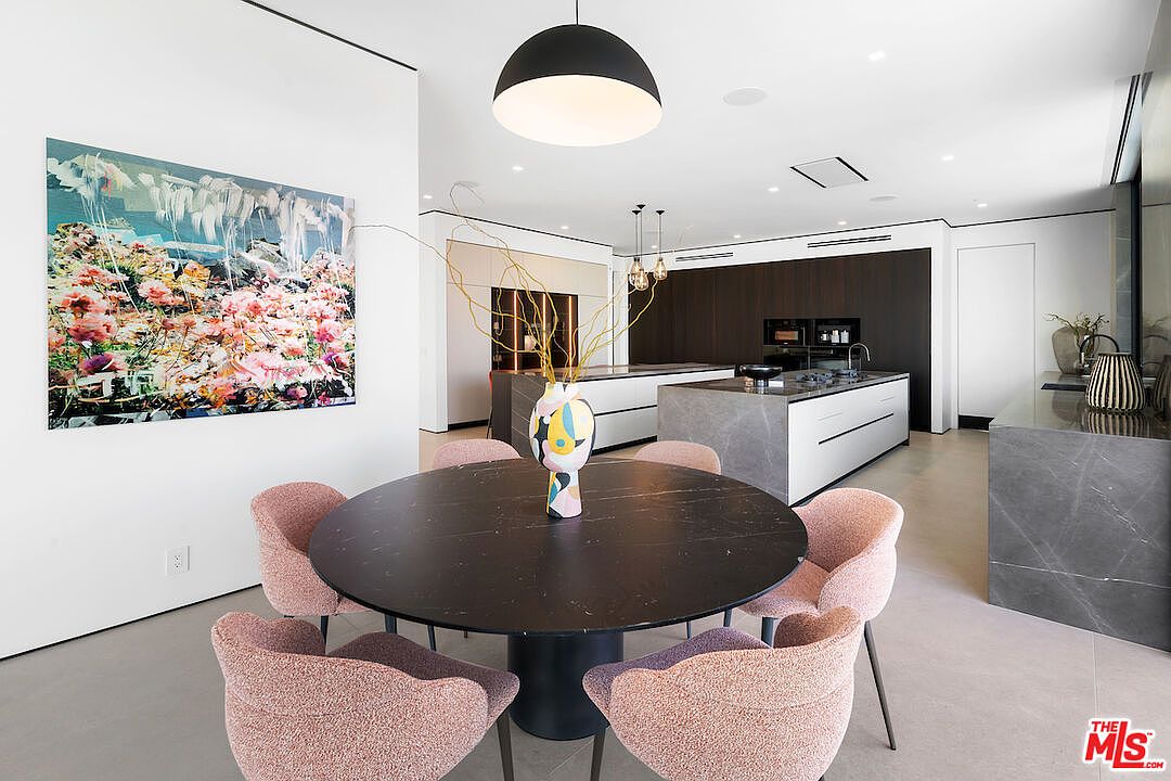 This open-concept dining area features a circular dark stone table surrounded by four textured pink chairs, positioned beneath a large, modern black dome pendant light. The space flows seamlessly into a contemporary kitchen equipped with a large stone-topped island and dark cabinetry. A vibrant, abstract floral painting adds a bold splash of color to the minimalist white wall, creating a sophisticated and artistic atmosphere.