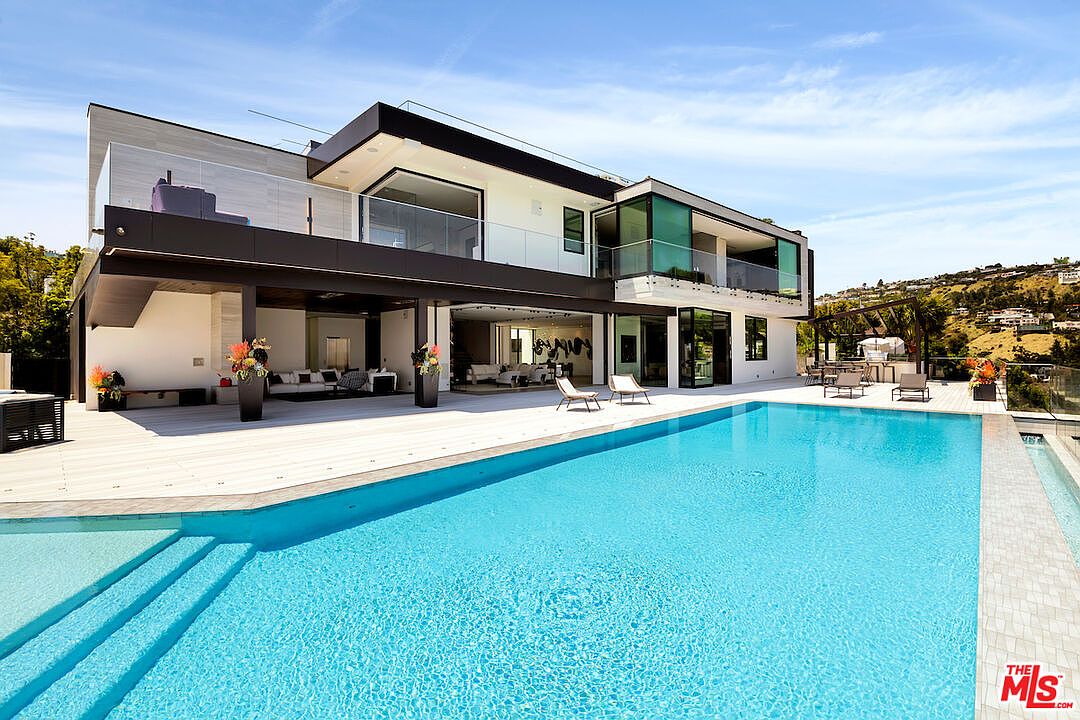 This modern luxury residence features a sprawling, multi-level architectural design with a large, rectangular swimming pool as the focal point of the backyard. The home showcases expansive glass walls that open to a covered patio area, creating a seamless transition between indoor and outdoor living spaces. The scene is set under a bright blue sky, highlighting the clean lines, neutral exterior finishes, and the inviting, resort-style atmosphere of the property.