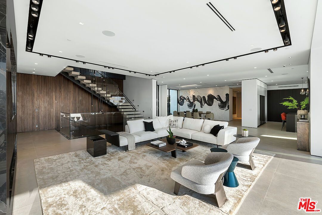 This expansive, open-concept living area features a modern aesthetic with a large white sectional sofa, two textured armchairs, and a low-profile coffee table centered on a neutral area rug. A striking staircase with glass railings and dark wood paneling serves as a focal point, while recessed lighting and a linear track lighting system illuminate the space. The room flows seamlessly into a dining area in the background, creating a bright and airy atmosphere suitable for sophisticated entertaining.