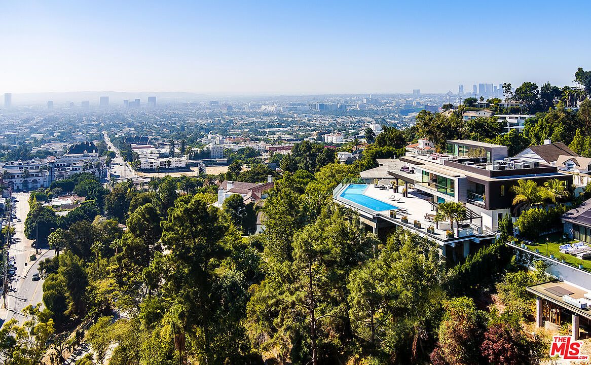 This high-angle aerial shot captures a luxurious modern residence perched on a hillside, overlooking a sprawling cityscape. The home features a sleek, multi-level architectural design with a prominent infinity-edge pool extending over the edge of the terrace, offering panoramic views. Lush greenery surrounds the property, emphasizing its secluded and elevated position within the landscape.