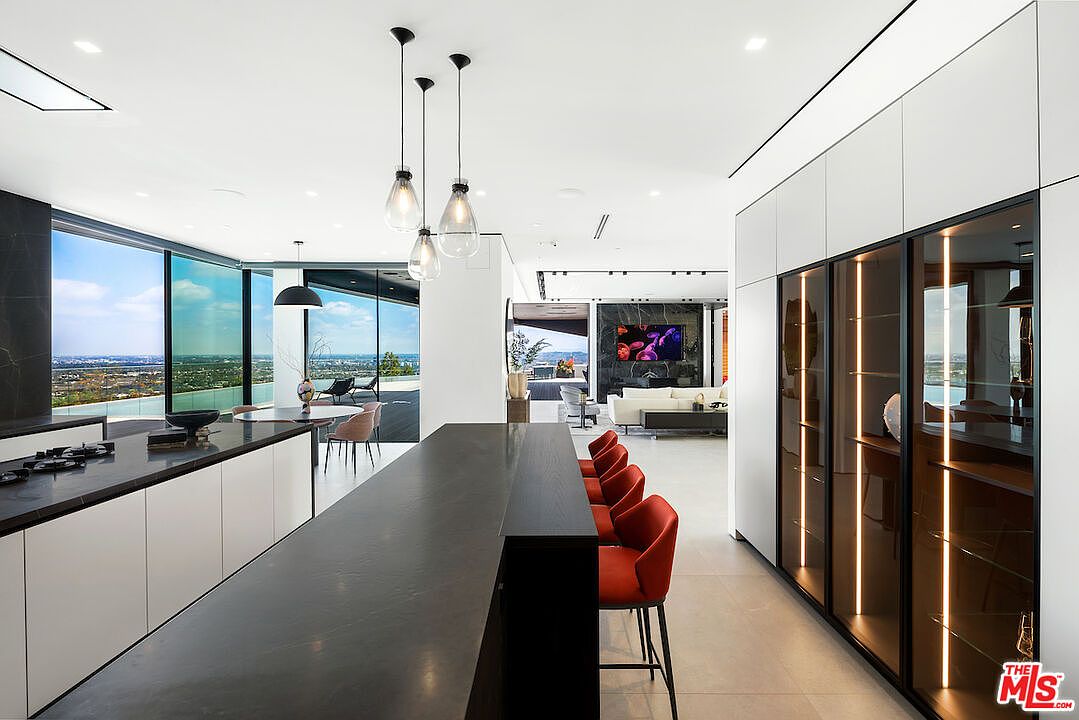 This modern, open-concept kitchen features a long, dark-toned island with integrated seating and a row of vibrant red bar stools. To the right, a wall of sleek, white cabinetry is complemented by tall, glass-fronted display cabinets with integrated vertical lighting. The space flows seamlessly into a bright living area with large glass walls that provide an expansive, panoramic view of the city skyline.
