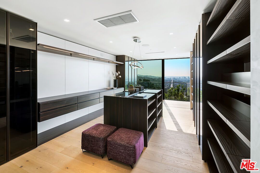 This luxurious walk-in closet features dark wood cabinetry, a central island with storage, and two plush purple ottomans. The space is illuminated by recessed lighting and a modern pendant fixture, while floor-to-ceiling glass windows offer an expansive view of the city skyline. The light wood flooring and clean, minimalist design create a sophisticated and organized atmosphere.