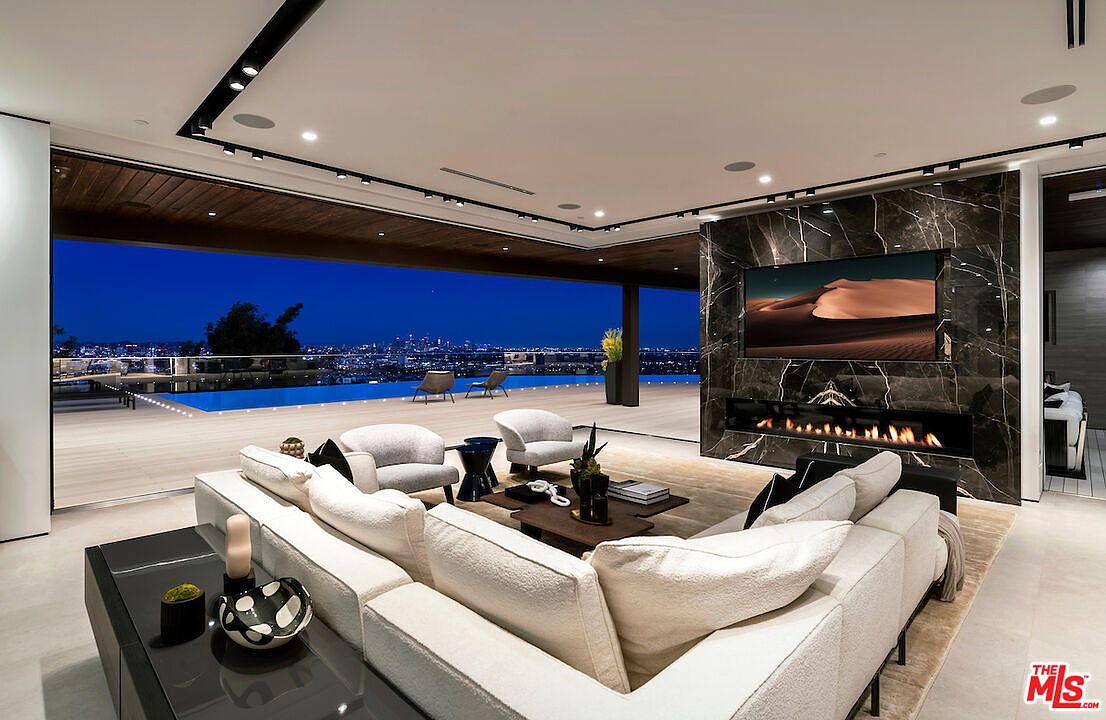 This expansive, modern living room features an open-concept design that seamlessly transitions to an outdoor deck through large, retractable glass walls. The space is anchored by a dark, stone-clad chimney breast housing a wide, linear fireplace and a mounted television. A plush, light-colored sectional sofa and several accent chairs create a comfortable seating area, while the backdrop showcases a stunning panoramic view of a city skyline at dusk.