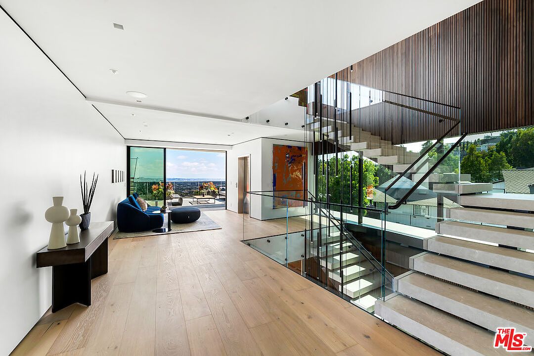 This modern, open-concept interior space features a striking floating staircase with glass railings and a dramatic light fixture installation. The area flows seamlessly into a living space with large sliding glass doors that open to an outdoor deck, showcasing expansive views. The design is characterized by light wood flooring, clean white walls, and a minimalist aesthetic that emphasizes architectural transparency.