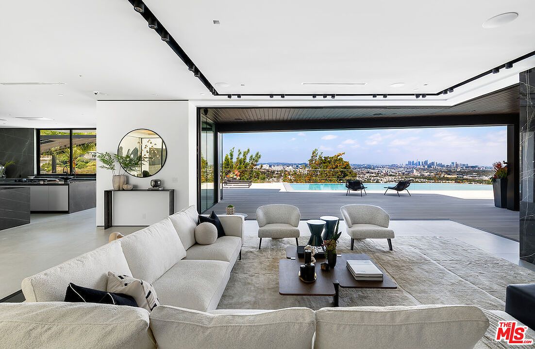 This expansive, modern living room features an open-concept design that seamlessly transitions to an outdoor deck through large, retractable glass walls. The space is furnished with a plush, light-toned sectional sofa and two accent chairs arranged around a tiered coffee table, all set upon a neutral area rug. The room is illuminated by recessed track lighting and offers a panoramic view of the city skyline and a sparkling infinity-edge pool.