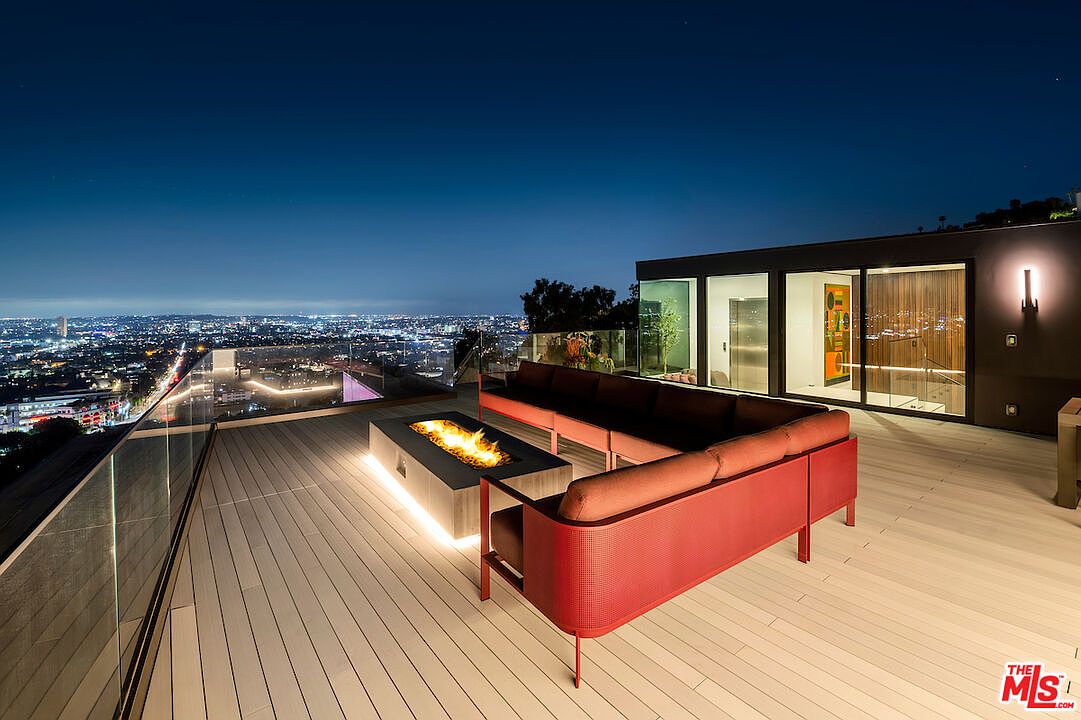 This modern rooftop deck offers a panoramic view of the city skyline at night, featuring a sleek, low-profile red sectional sofa arranged around a rectangular fire pit. The deck is constructed with light-toned composite decking and is enclosed by a frameless glass railing, creating an unobstructed connection to the urban landscape. Large glass sliding doors provide a seamless transition from the outdoor lounge area to the interior living space.