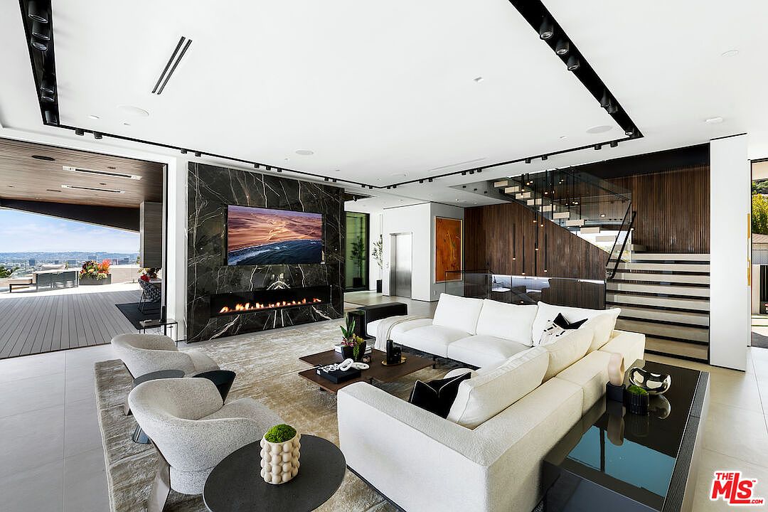 This expansive, modern living room features a striking black stone-clad chimney breast that serves as a focal point for the integrated television and linear fireplace. The space is furnished with a large, plush white sectional sofa and contemporary armchairs, all arranged on a textured area rug. Large sliding glass doors open to an outdoor deck, creating a seamless transition between the indoor living area and the exterior space, while a staircase with glass railings adds a sophisticated architectural element to the room.