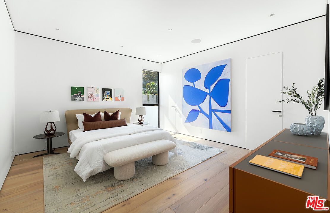 This modern primary bedroom features a neutral color palette with light wood flooring and crisp white walls. A large, abstract blue art piece serves as a focal point, complemented by a plush bed with a textured bench at its foot and a series of smaller framed artworks above the headboard. The room is bright and airy, with a window providing natural light and a sleek dresser positioned in the foreground.