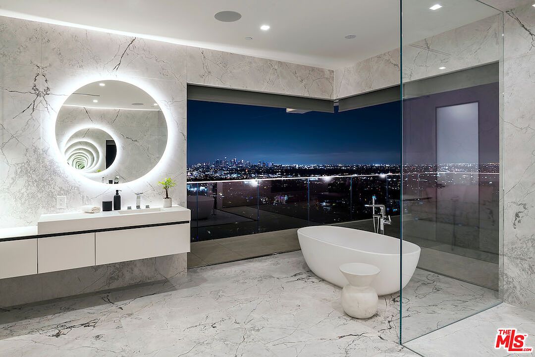This luxurious primary bathroom features a sleek, wall-mounted vanity with a circular backlit mirror and a freestanding soaking tub positioned near expansive glass walls. The room is finished with light-toned, veined stone surfaces throughout the walls and flooring, creating a cohesive and high-end aesthetic. A stunning, unobstructed nighttime view of a sprawling city skyline serves as the dramatic backdrop for the space.