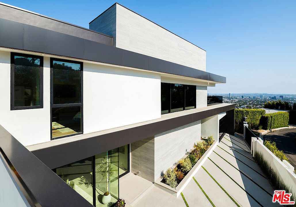This modern architectural residence features a striking geometric design with clean white walls, dark horizontal trim, and large glass windows. The perspective is elevated, looking down at a paved driveway with integrated grass strips and a landscaped planter bed filled with shrubs. The home offers a panoramic view of the distant city skyline, emphasizing its elevated hillside location.