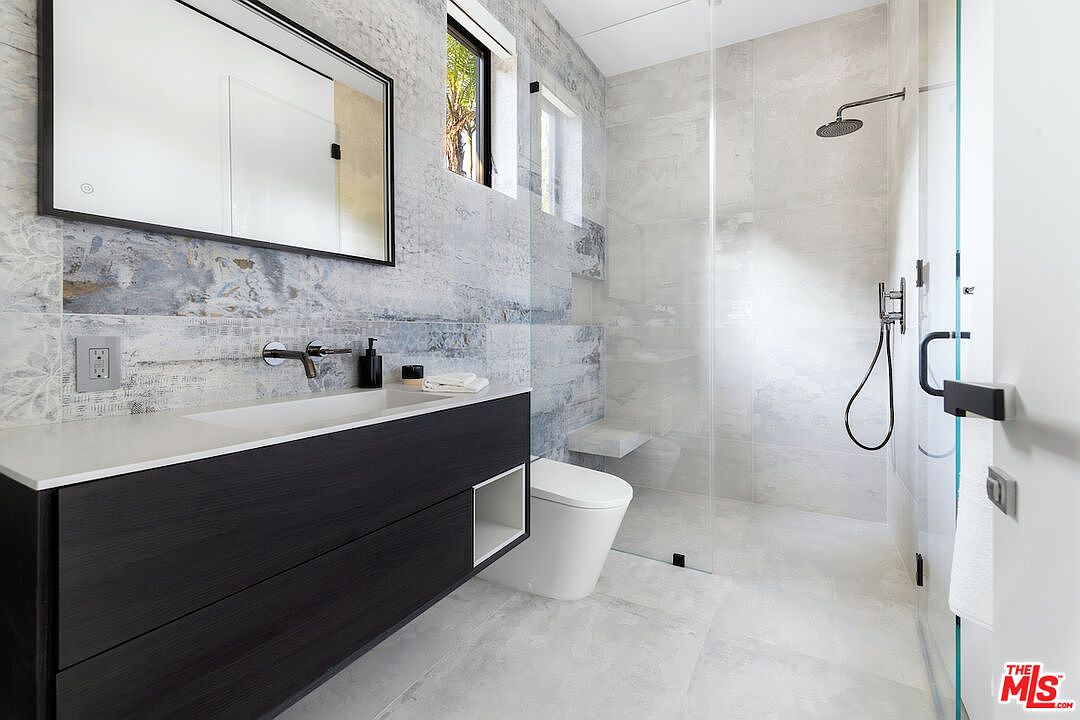 This modern bathroom features a floating vanity with a dark wood finish and a white countertop, paired with a sleek rectangular mirror. The walls are adorned with textured, multi-toned stone tiles that extend into the walk-in shower area, which is enclosed by a glass partition. A wall-mounted toilet and minimalist fixtures complete the contemporary, high-end aesthetic of the space.