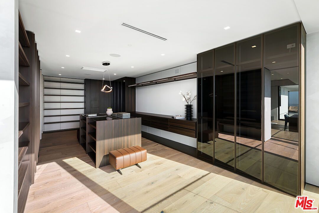This spacious, modern walk-in closet features a central island with dark wood cabinetry and a sleek countertop, complemented by a leather-upholstered bench. The room is lined with extensive storage, including open shelving on the left and large, dark-tinted glass-front wardrobes on the right. Light wood flooring runs throughout the space, which is illuminated by recessed ceiling lights and a contemporary pendant fixture.