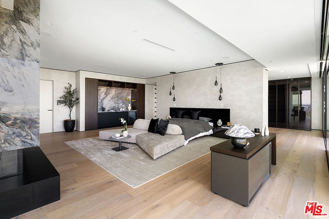 This expansive primary bedroom features a contemporary design with light wood flooring and a neutral color palette. A large, low-profile bed is positioned in the center of the room, complemented by a modern sectional sofa and a sleek wet bar area with dark cabinetry. The space is illuminated by unique pendant lighting and offers a sophisticated, open-concept layout with a view into a glass-enclosed closet area.