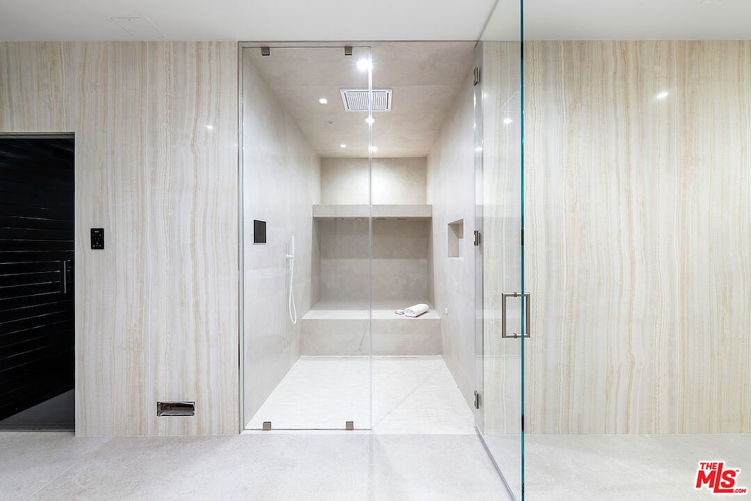 This modern, minimalist shower space features a large, walk-in enclosure with floor-to-ceiling glass panels. The interior is finished in neutral, light-toned stone tiles and includes a built-in bench, a recessed wall niche, and a rainfall showerhead. To the left, a dark, textured door provides a striking contrast to the light, vertical-veined stone wall cladding the surrounding area.