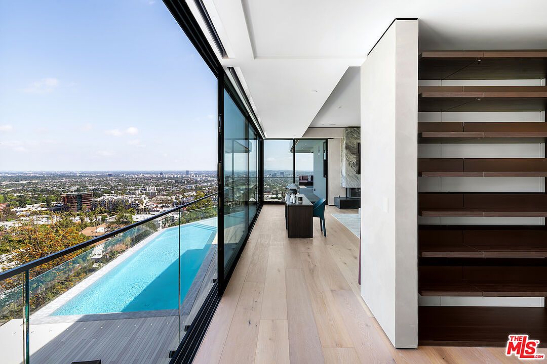 This modern interior space features a sleek desk positioned alongside expansive floor-to-ceiling glass walls that offer a panoramic view of the city and an adjacent infinity-edge pool. To the right, a set of open-tread wooden stairs adds architectural interest, while light wood flooring runs throughout the open-concept area. The perspective captures a seamless transition between the refined indoor workspace and the dramatic outdoor landscape.