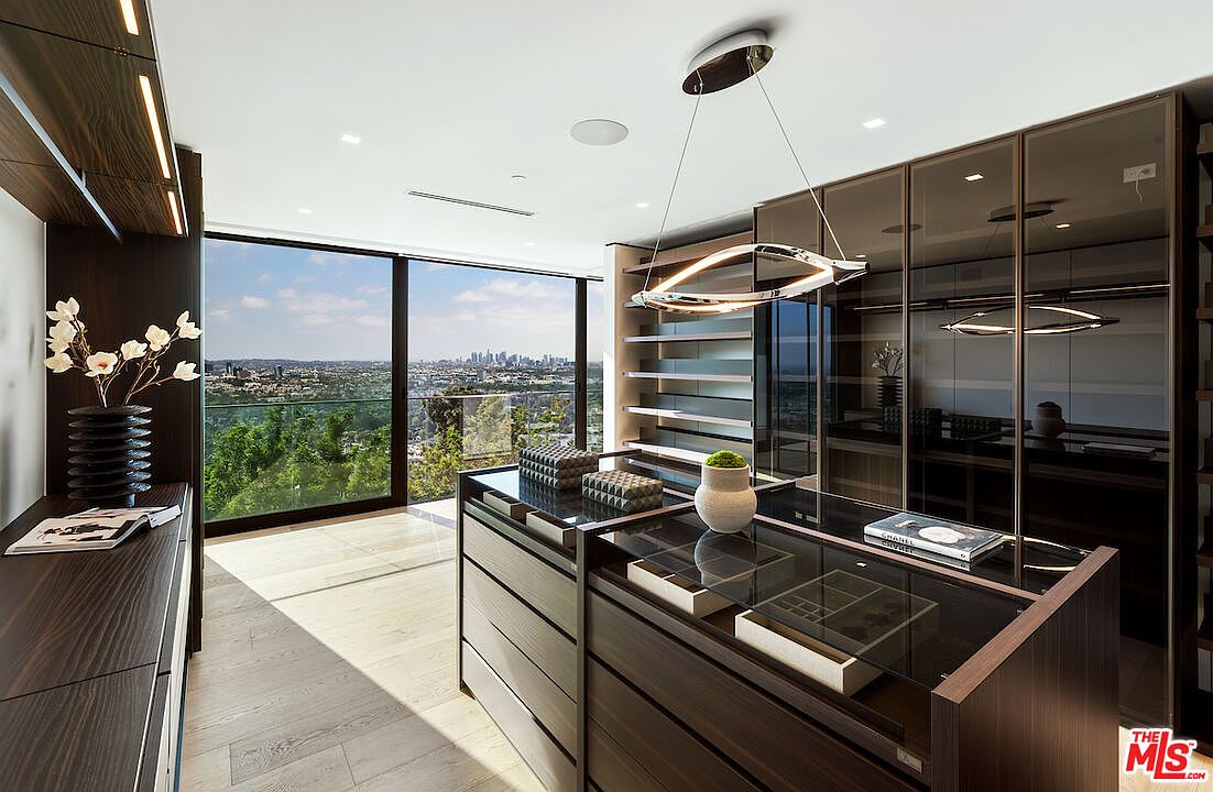 This luxurious walk-in closet features dark wood cabinetry with glass-fronted doors and a central island topped with a glass surface for displaying accessories. A modern, sculptural light fixture hangs above the island, while floor-to-ceiling windows provide an expansive, panoramic view of the city skyline and surrounding hills. The space is bright and airy, combining high-end storage solutions with a sophisticated, contemporary aesthetic.