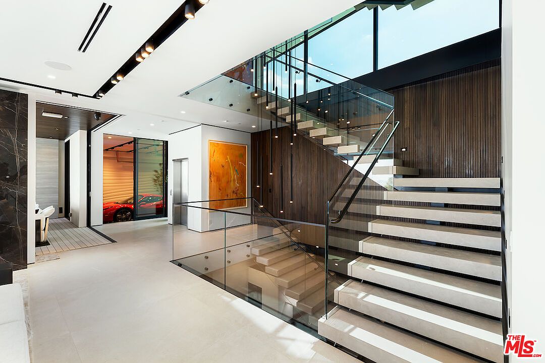 This grand, modern entryway features a striking floating staircase with light-toned treads and a glass railing that overlooks the lower level. A dramatic wall of dark, vertical wood slats serves as a backdrop for a series of slender, hanging pendant lights, while a vibrant abstract painting adds a pop of color to the minimalist space. To the left, a glass-enclosed garage display showcases a red sports car, emphasizing the home's luxurious and contemporary aesthetic.