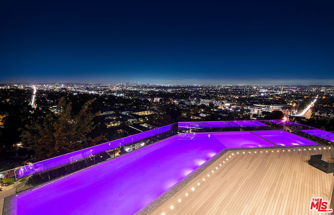 This elevated perspective captures a stunning infinity-edge pool illuminated with vibrant purple lighting, set against a panoramic night view of the sprawling city skyline. A sleek wooden deck borders the pool, featuring a modern glass railing that ensures an unobstructed view of the urban lights below. The composition emphasizes a luxurious, high-end outdoor living space designed for evening entertainment and relaxation.