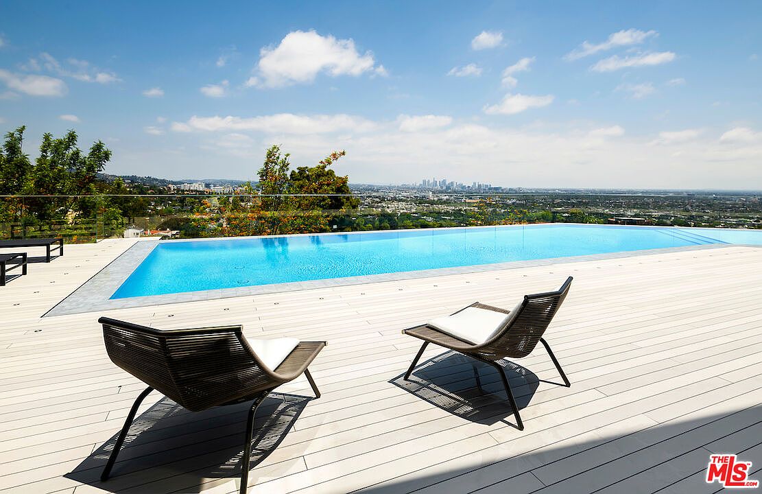 This image captures a sleek, modern infinity-edge pool set against a sprawling panoramic view of the city skyline. The foreground features a light-toned wooden deck furnished with two contemporary lounge chairs, creating a serene outdoor retreat. The composition emphasizes the seamless transition between the pool's water and the distant horizon, highlighting the property's elevated and luxurious setting.