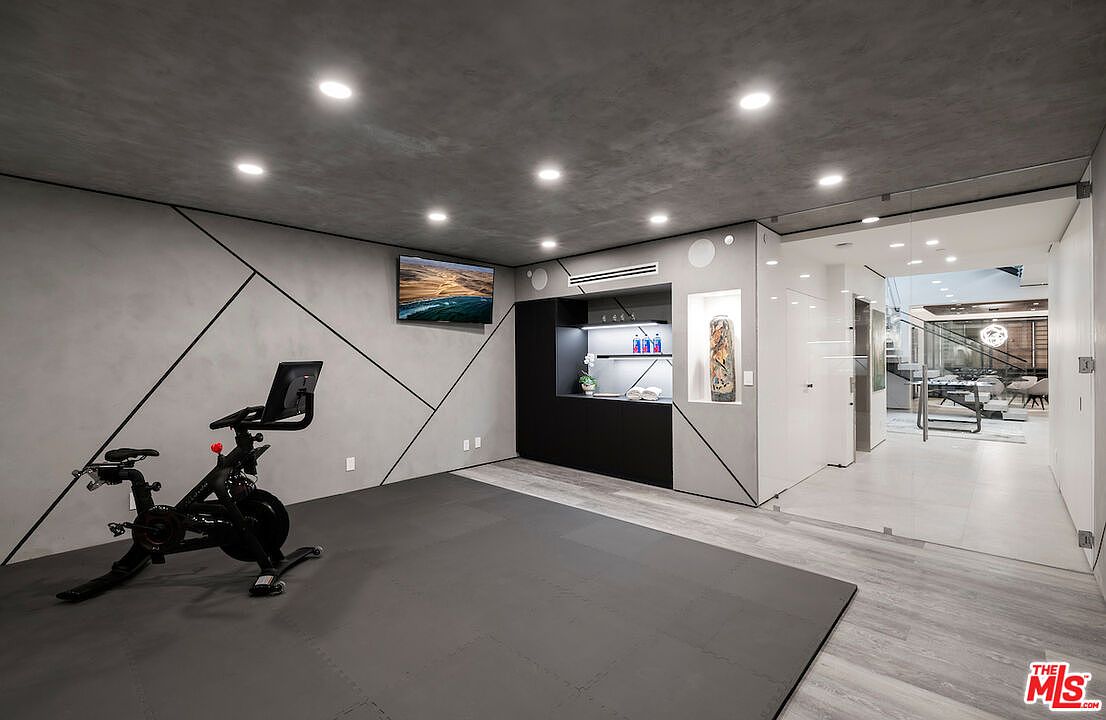 This modern home gym features a sleek, minimalist aesthetic with gray walls accented by geometric black lines and a matching gray ceiling. A stationary exercise bike sits on a dark rubberized mat, while a built-in black cabinetry unit provides storage and a small refreshment area. The space is illuminated by recessed lighting and connects to an adjacent living area through a large glass partition, creating an open and airy feel.