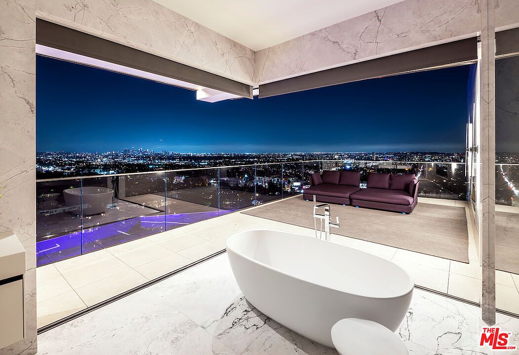 This luxurious primary bathroom features a sleek, freestanding soaking tub positioned on a light-toned stone floor. The space is defined by expansive, floor-to-ceiling glass walls that open onto a private balcony, offering a panoramic view of the city skyline at night. A plush, dark-toned sofa sits on the balcony, creating a sophisticated indoor-outdoor living experience.