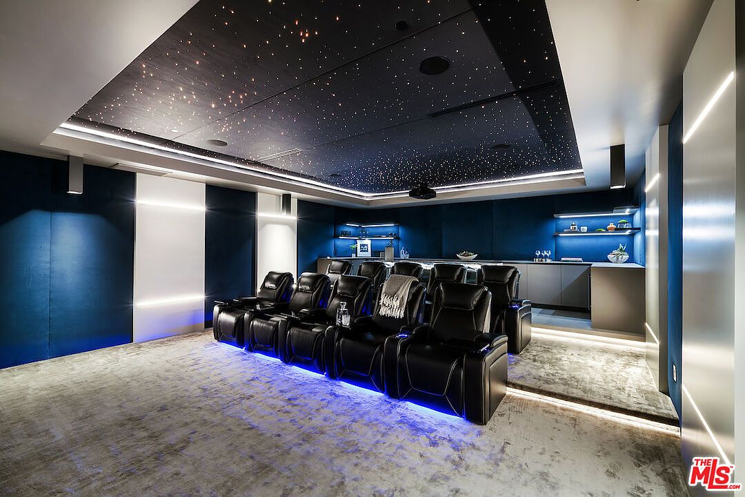 This image showcases a luxurious, modern home theater room designed for an immersive cinematic experience. The space features a row of plush, black leather reclining seats illuminated by blue LED floor lighting, set against a backdrop of dark blue acoustic wall panels and a dramatic starlit ceiling. A built-in refreshment bar with illuminated shelving and cabinetry is positioned at the rear, completing the sophisticated and high-end entertainment environment.
