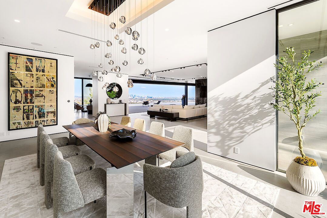 This sophisticated dining area features a long, dark-toned table surrounded by textured, light-colored upholstered chairs, set beneath a striking cluster of spherical pendant lights. The open-concept space flows seamlessly into a bright living area with expansive glass walls that reveal a panoramic city view. A large, abstract art piece adorns the wall, while a potted tree adds a touch of organic texture to the minimalist, modern interior.