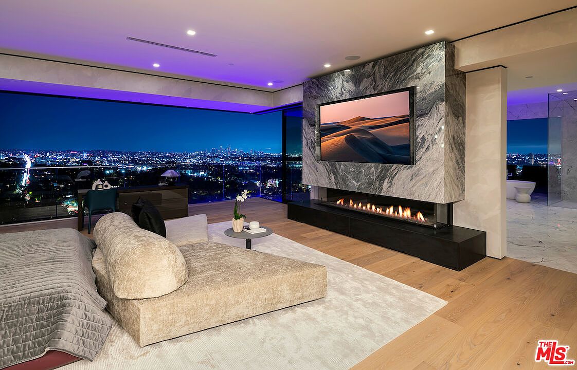This luxurious primary bedroom features a contemporary design with a large, plush bed and a sleek, low-profile lounge chair. A prominent stone-clad chimney breast houses a wide, linear fireplace beneath a wall-mounted television, creating a sophisticated focal point. The room is defined by expansive glass walls that offer panoramic nighttime city views, complemented by recessed purple accent lighting and light-toned wood flooring.