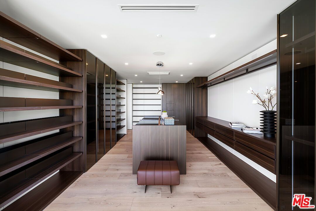This expansive, modern walk-in closet features dark wood cabinetry with a mix of open shelving and glass-fronted doors. A central island provides additional storage and surface space, complemented by a leather-upholstered bench, while light wood flooring runs throughout the room. The space is illuminated by recessed lighting and a sleek pendant fixture, creating a sophisticated and organized atmosphere.
