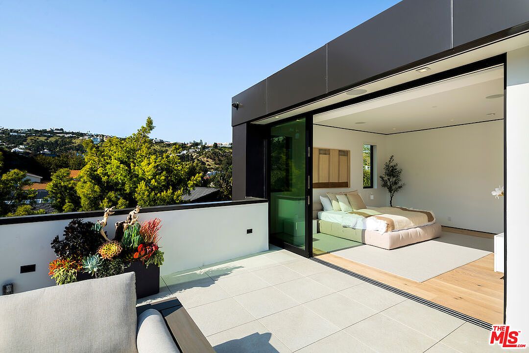 This image captures a modern balcony featuring light-colored stone tiles and a sleek black metal railing that overlooks a scenic hillside landscape. A large sliding glass door connects the outdoor space to a minimalist bedroom, creating a seamless indoor-outdoor living experience. The balcony is accented by a large planter filled with vibrant succulents, enhancing the contemporary aesthetic of the home.