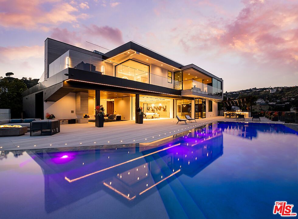 This stunning contemporary residence features a sprawling deck that seamlessly integrates with a large, illuminated infinity-edge pool. The architecture showcases clean lines, expansive glass walls, and a sophisticated indoor-outdoor flow, all set against a dramatic sunset sky. The scene captures a luxurious lifestyle with outdoor lounge seating and a distant view of the hillside landscape.