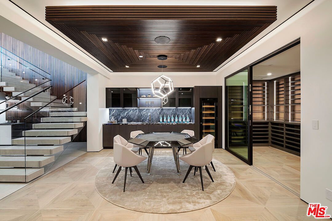 This sophisticated game room features a central octagonal table surrounded by four modern, light-toned chairs, all set upon a circular area rug. The space is defined by a striking dark wood-slatted ceiling with recessed lighting and a geometric pendant fixture, while a wet bar with dark cabinetry and a stone backsplash sits against the back wall. To the left, a floating staircase with glass railings adds architectural interest, and to the right, a glass-enclosed wine storage area completes the luxurious, contemporary aesthetic.