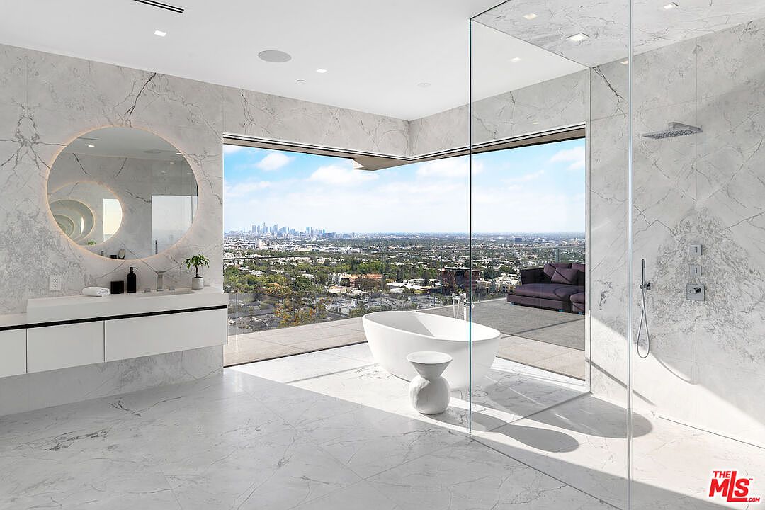 This luxurious primary bathroom features expansive floor-to-ceiling glass walls that provide an unobstructed, panoramic view of the city skyline. The space is finished with light-toned stone surfaces, a sleek floating vanity with a circular backlit mirror, and a freestanding soaking tub positioned to take advantage of the vista. A glass-enclosed shower area completes the minimalist, high-end aesthetic, creating a seamless transition between the indoor sanctuary and the outdoor balcony.