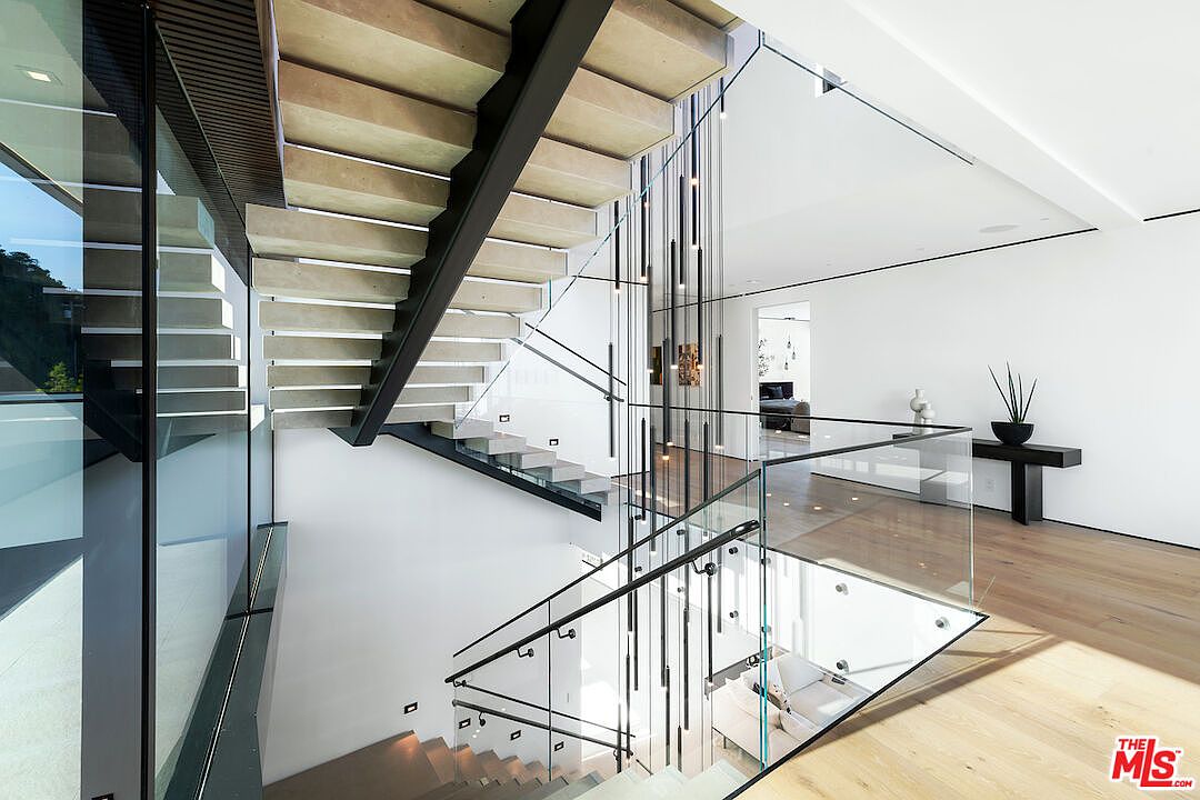 This striking interior view showcases a modern floating staircase with light wood treads and a black metal frame, set against a crisp white wall. A dramatic, multi-pendant light fixture hangs vertically through the stairwell, creating a sense of height and architectural sophistication. The space features glass railings and light wood flooring, leading to a bright landing area with a minimalist console table and decorative plant.