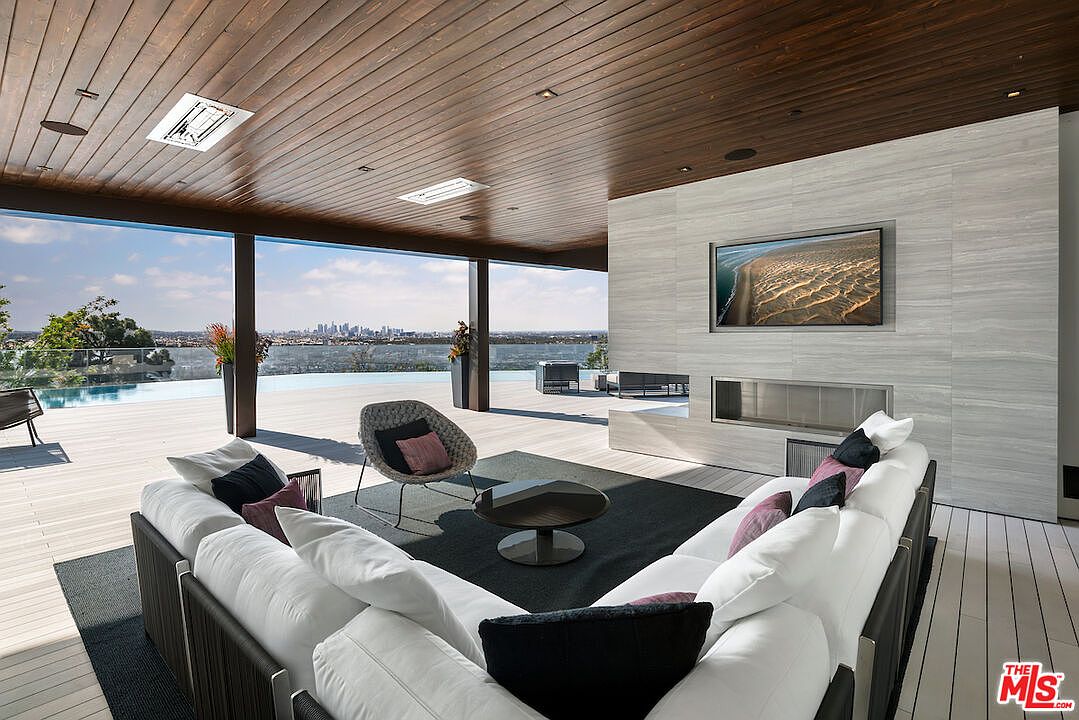 This expansive, covered outdoor living space features a modern aesthetic with a warm wood-paneled ceiling and a large, light-toned stone-clad wall housing a television and a linear fireplace. A plush, white sectional sofa and a circular accent chair are arranged on a dark area rug, creating a comfortable lounge area overlooking an infinity-edge pool and a panoramic city skyline. The open-air design seamlessly blends indoor comfort with the surrounding landscape, emphasizing a luxurious, resort-style atmosphere.