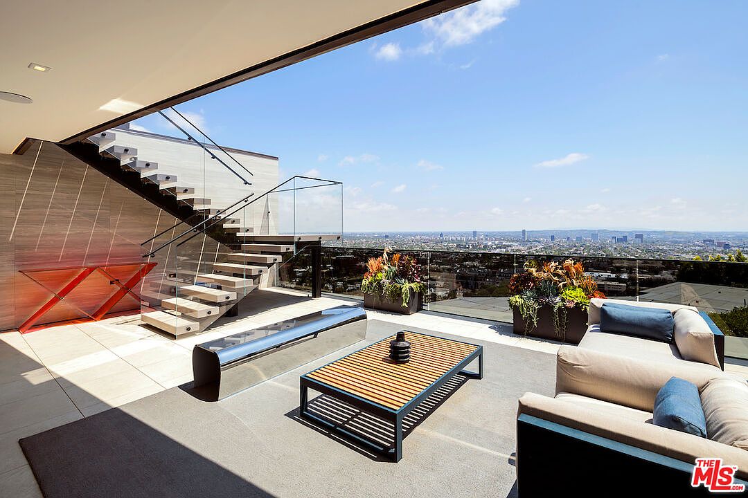 This expansive, modern outdoor terrace offers a panoramic view of the city skyline under a clear blue sky. The space features a sleek floating staircase with glass railings, a contemporary outdoor sofa, and a slatted wood coffee table, all set upon a neutral-toned patio surface. Large planters with lush greenery add a touch of nature to this sophisticated, open-air living area.